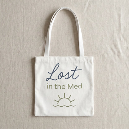 White tote bag with 'Lost in the Med' text and sun design on a beige background