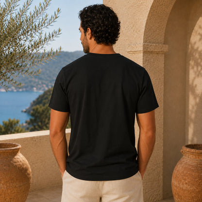 Man wearing a black t-shirt standing in front of a scenic view with mountains and water.
