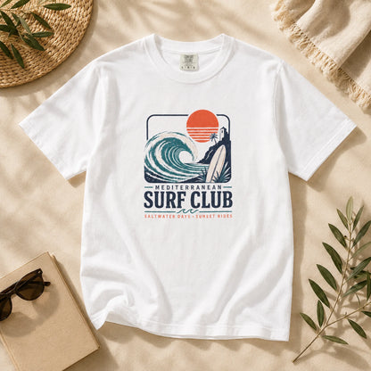 White t-shirt with 'Mediterranean Surf Club' graphic on a beige surface with sunglasses and a book.