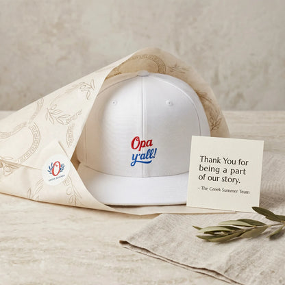 White cap with 'Opa y'all!' text, accompanied by a card and decorative elements on a neutral background