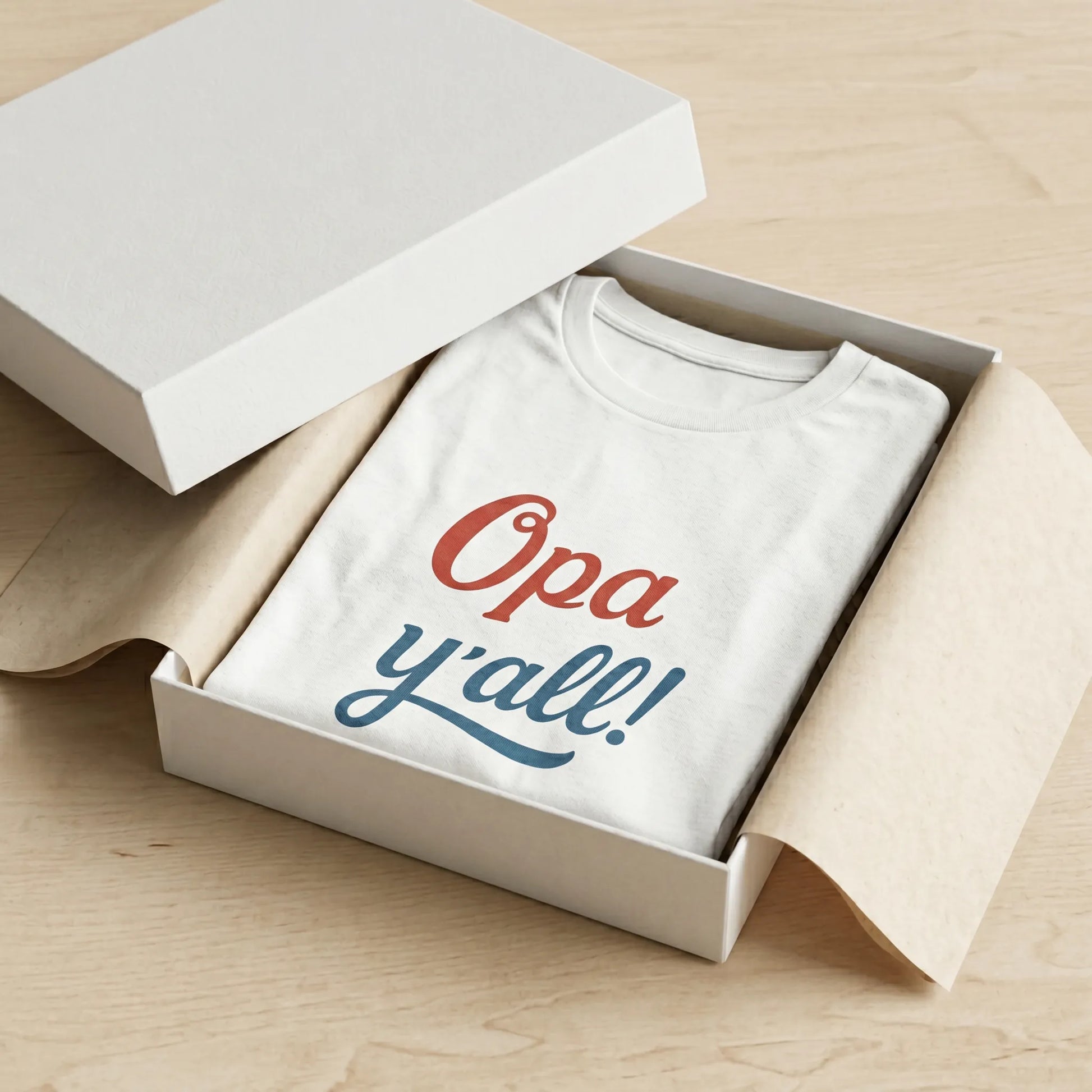 White t-shirt with 'Opa y'all!' text in red and blue inside an open box on a wooden surface.