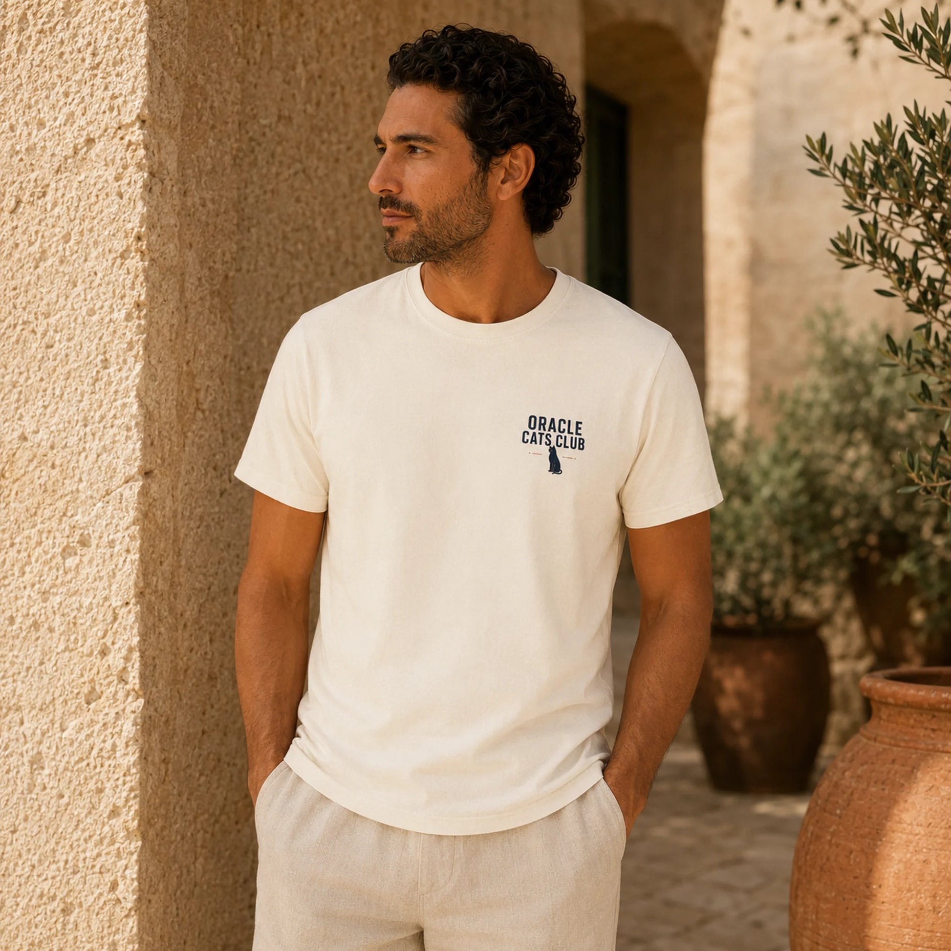 Man wearing a white t-shirt with a logo, standing outdoors against a beige wall.