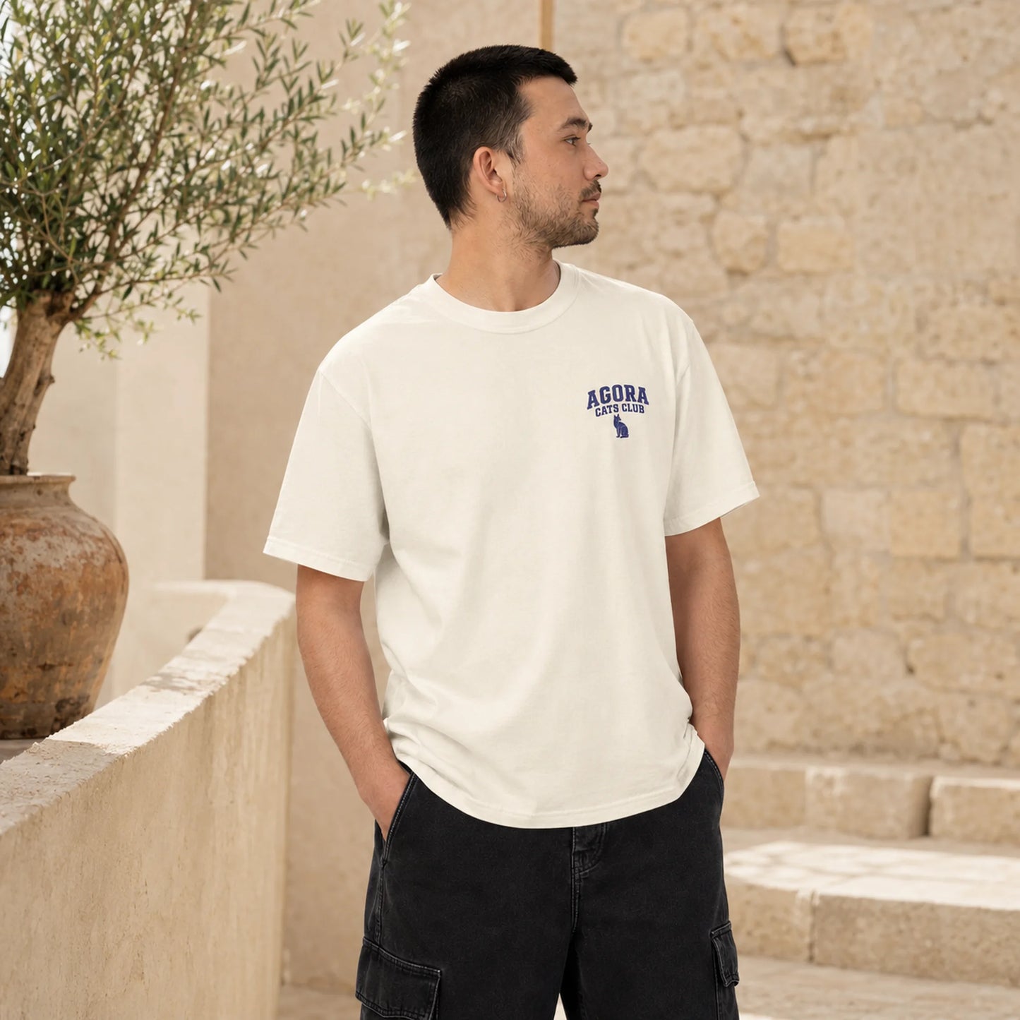 Man wearing a white t-shirt with 'Agora' logo outdoors against a stone wall.