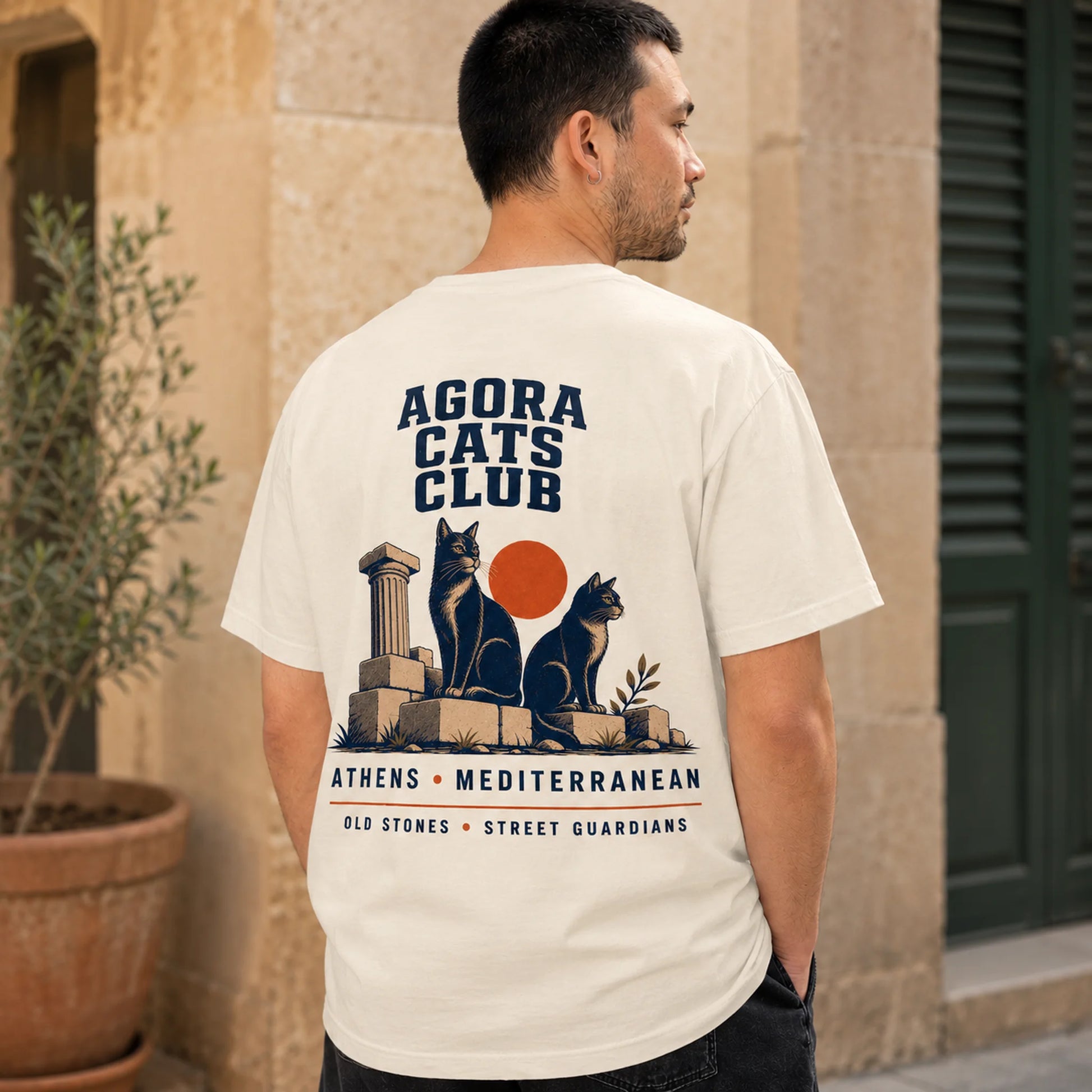 Man wearing a white t-shirt with 'Agora Cats Club' design, standing in front of a building.