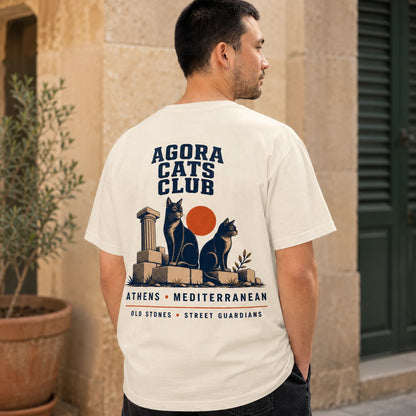 Man wearing a white t-shirt with 'Agora Cats Club' design, standing in front of a building.