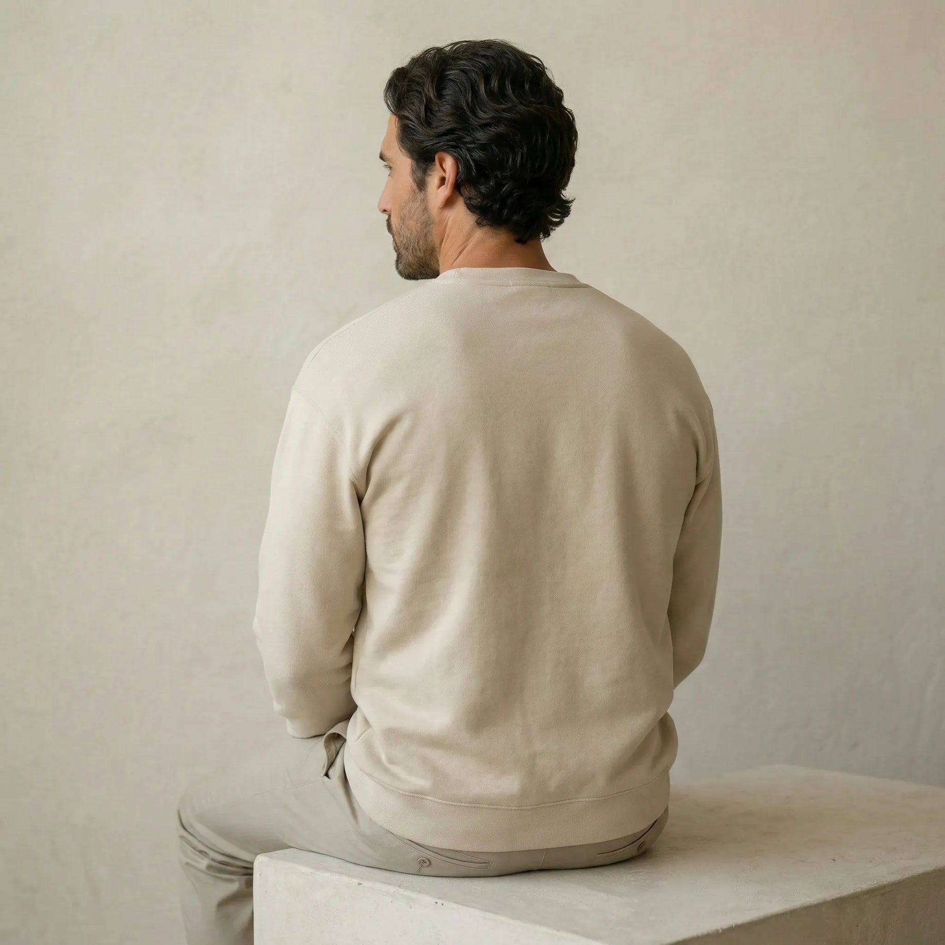 Man wearing a beige sweater sitting on a white surface against a plain wall.