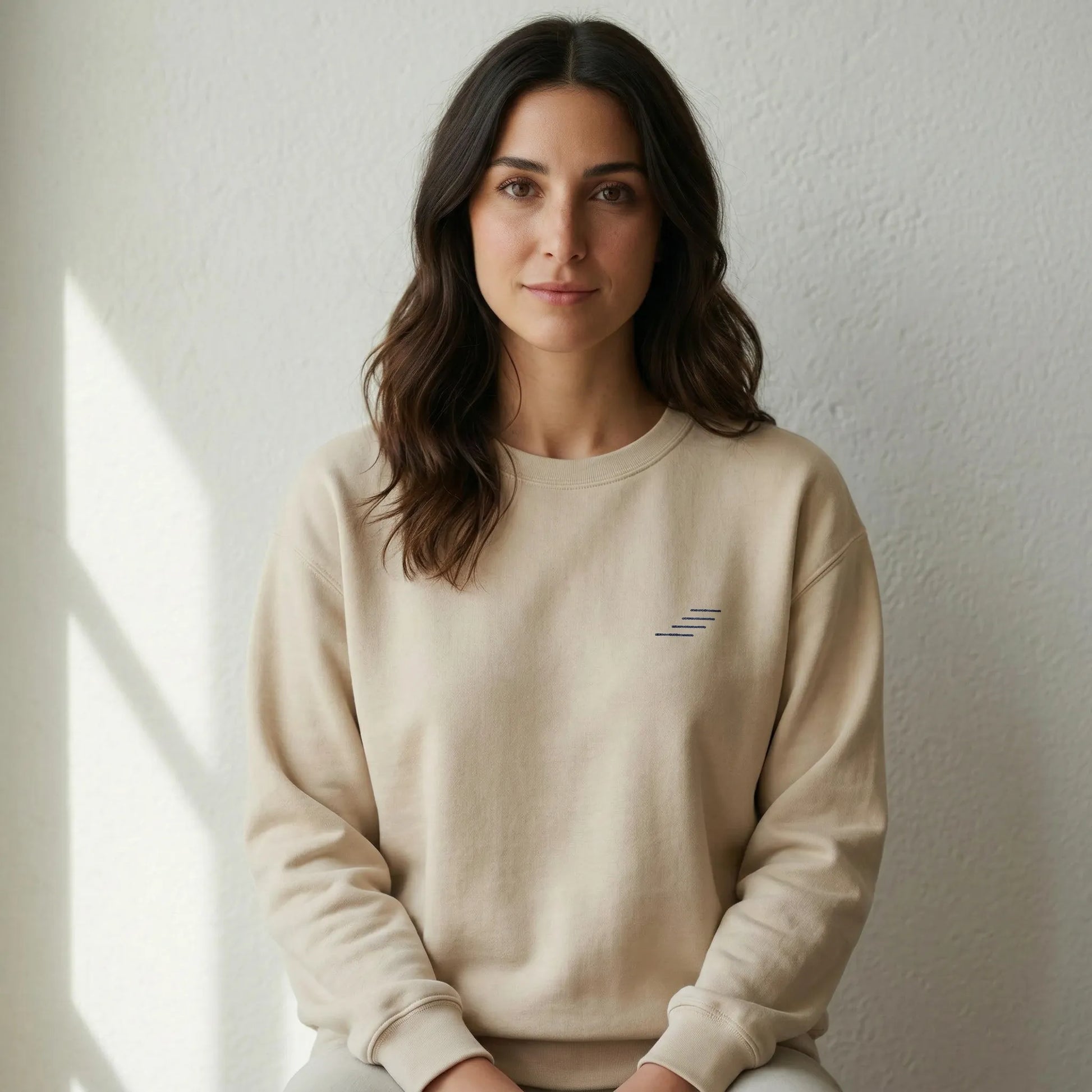 Woman wearing a beige sweatshirt with a small logo, sitting against a plain wall.