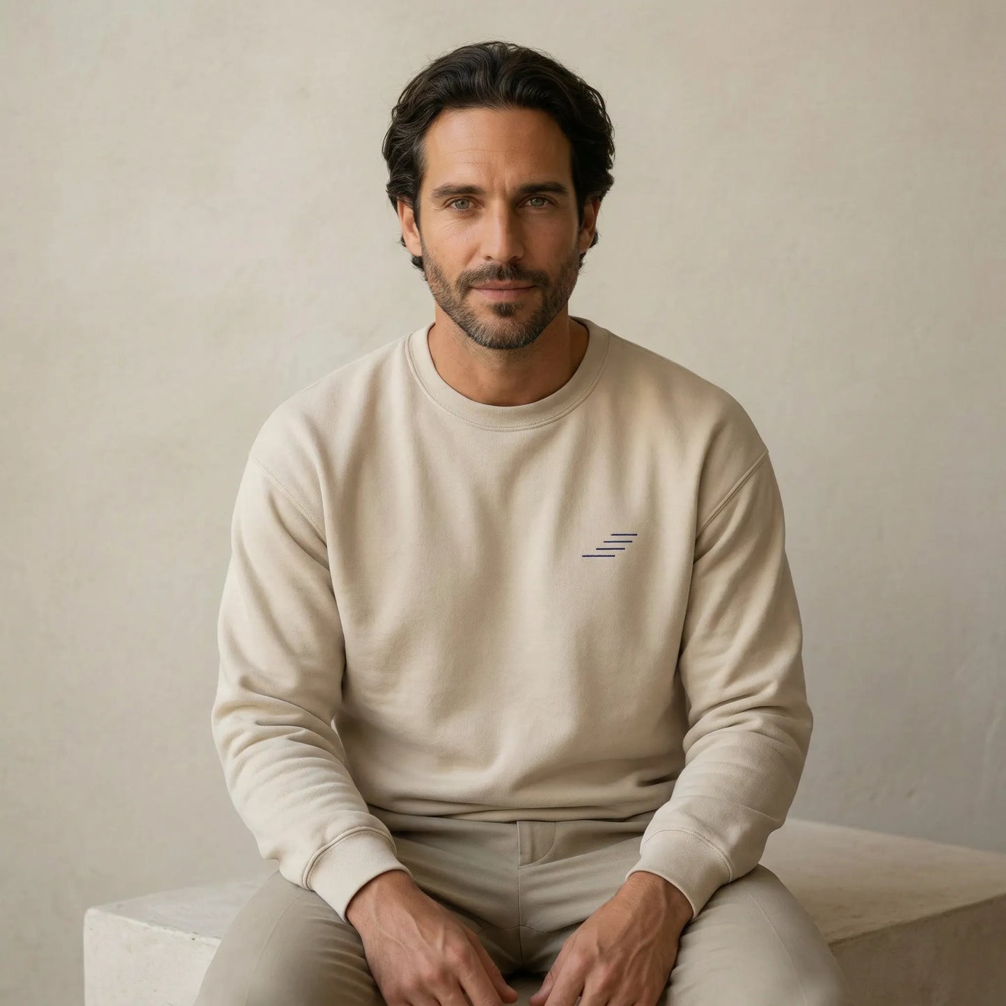 TP HERO: Man wearing a beige sweatshirt with a small logo, sitting on a light-colored surface against a neutral background.