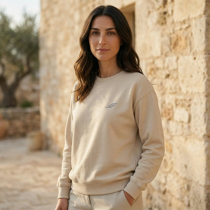 Woman wearing a beige sweatshirt with a small logo, standing in front of a stone wall.