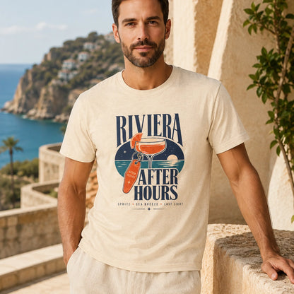 A man wearing a cream-colored graphic t-shirt with a retro-style 'Riviera After Hours' cocktail print, standing on a sunny stone balcony overlooking a scenic coastal landscape.
