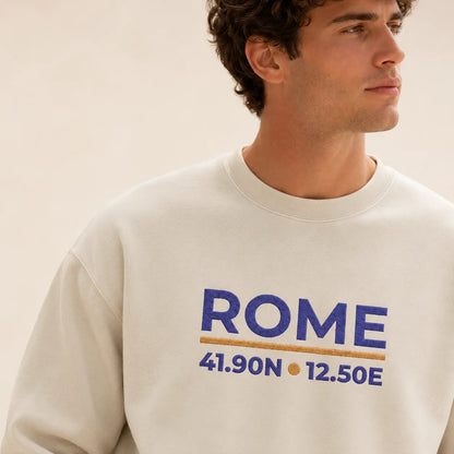 TP HERO: A man wearing a cream-colored crewneck sweatshirt with a blue and gold embroidered design that reads 'ROME 41.90N 12.50E'.