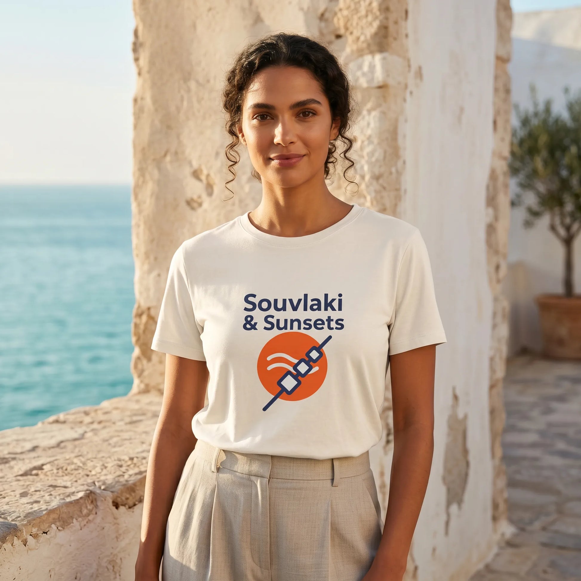 Woman wearing a 'Souvlaki & Sunsets' t-shirt by the sea.