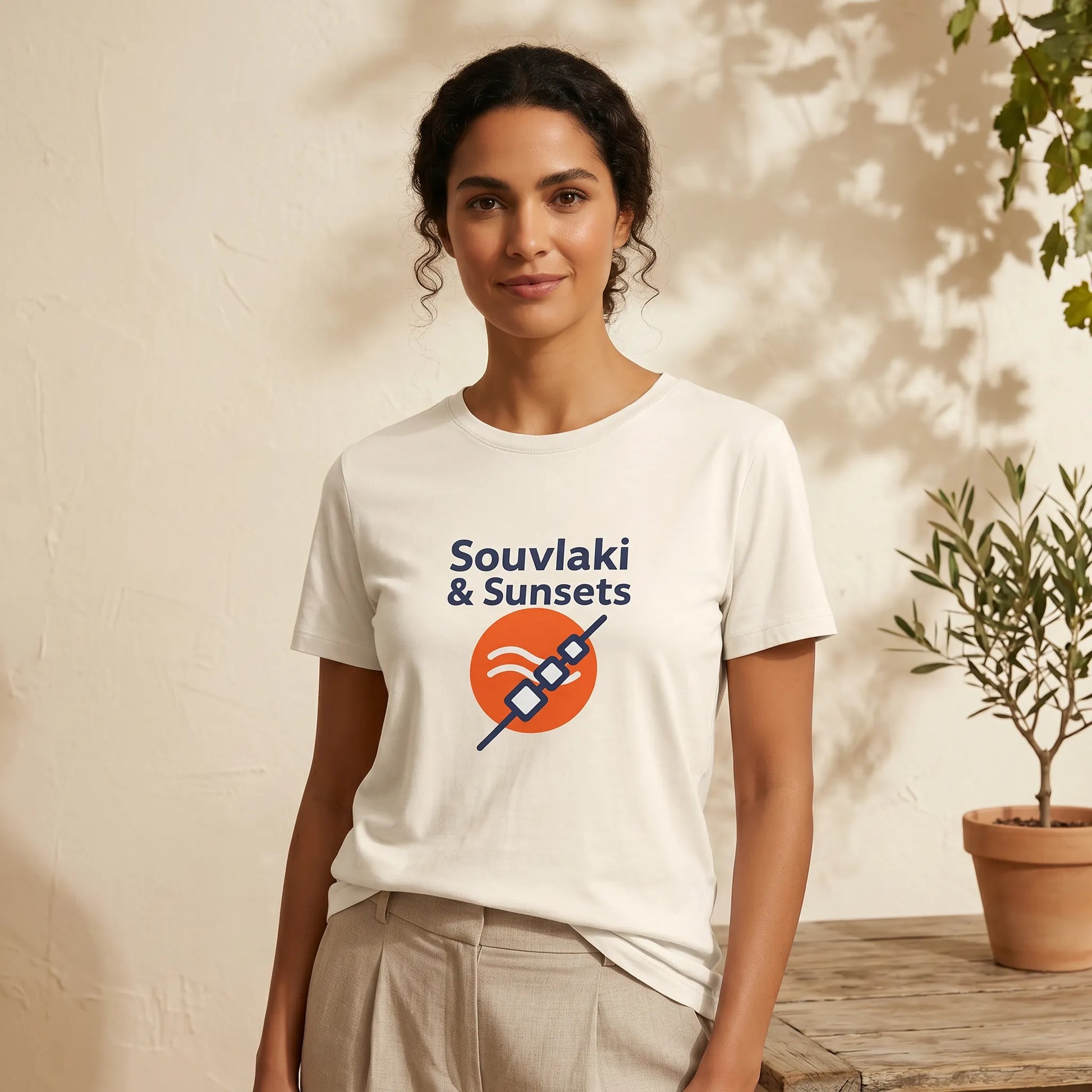 Woman wearing a white t-shirt with 'Souvaki & Sunsets' design, standing against a neutral background with a plant.