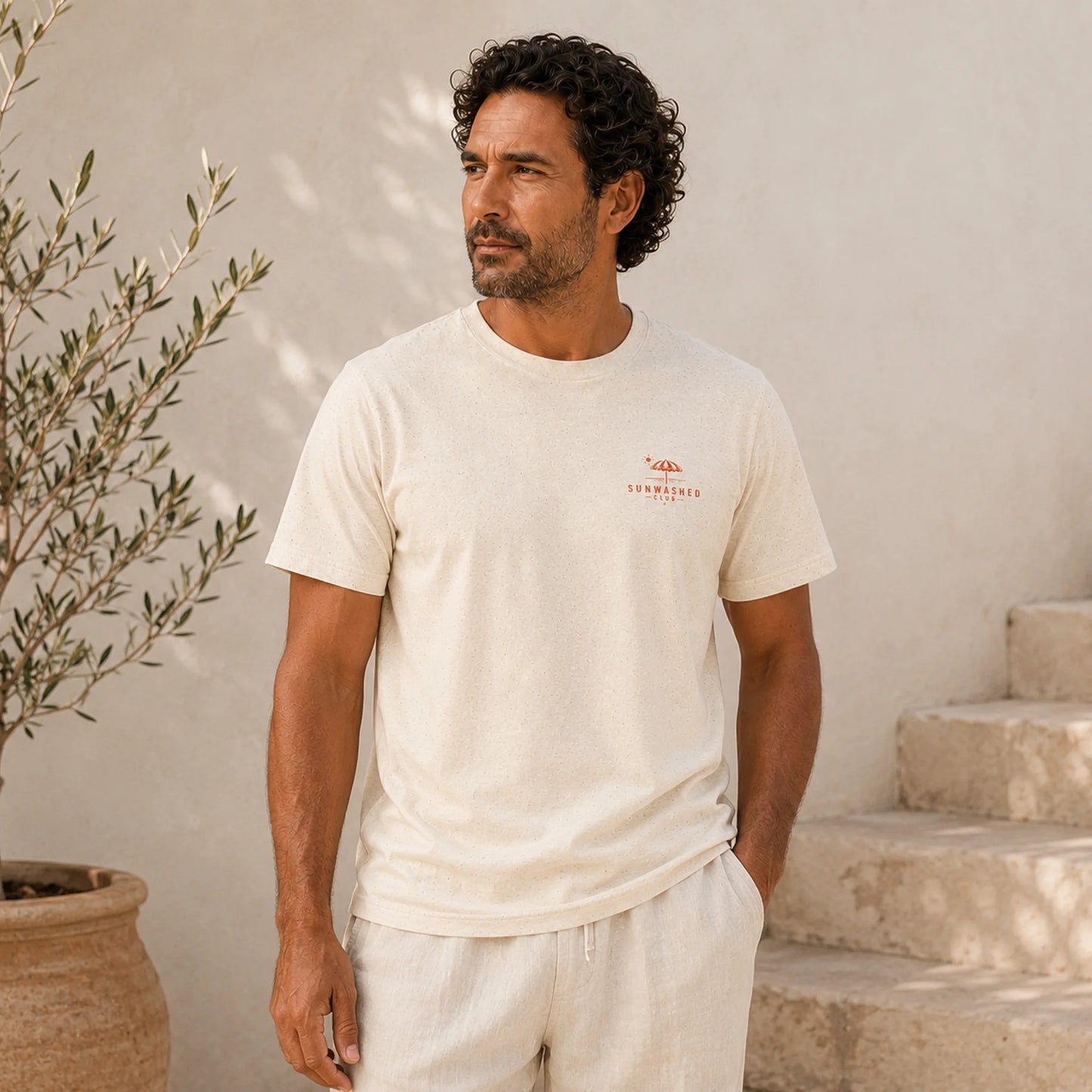 Man wearing a white t-shirt with a logo, standing outdoors against a light-colored wall.