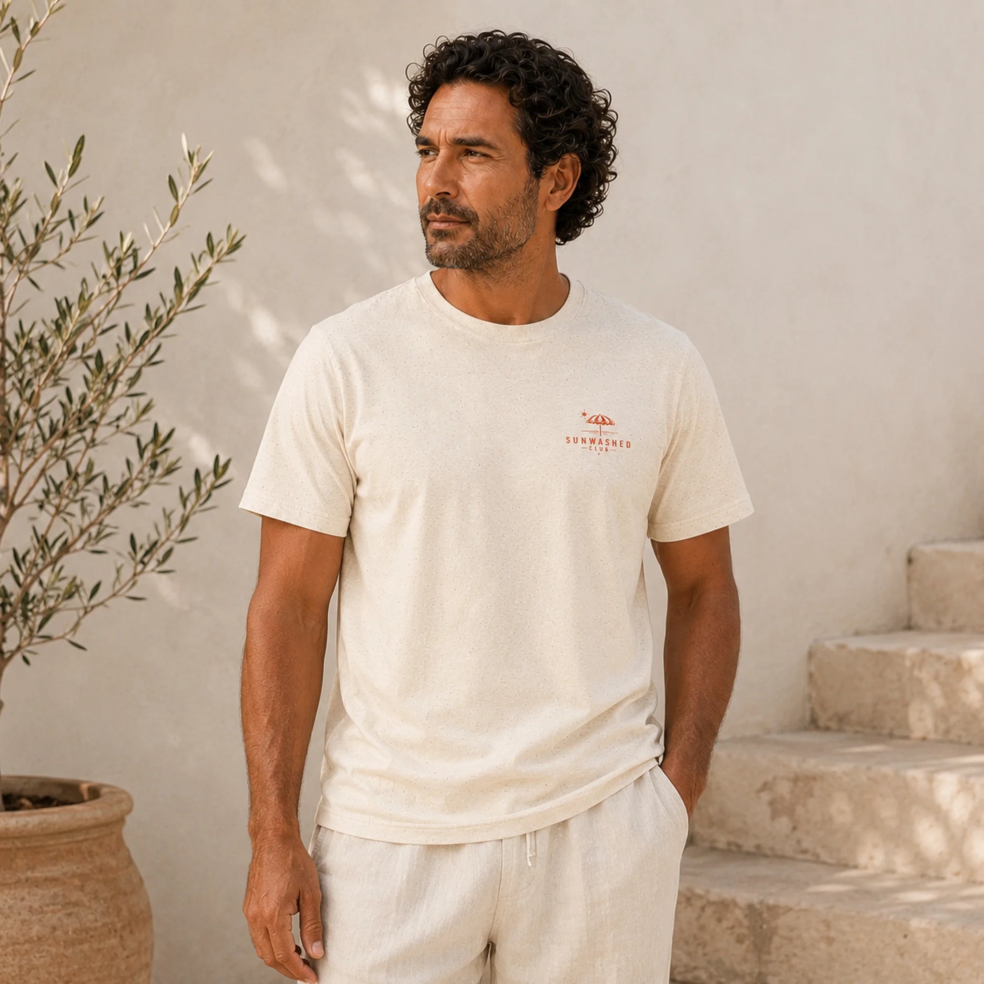 Man wearing a white t-shirt with a logo, standing outdoors against a light-colored wall.