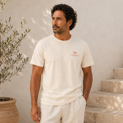 Man wearing a white t-shirt with a logo, standing outdoors against a light-colored wall.