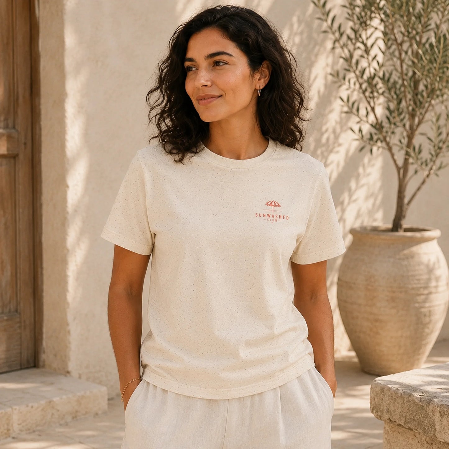 Woman wearing a beige t-shirt with a logo, standing outdoors near a building and potted plant.