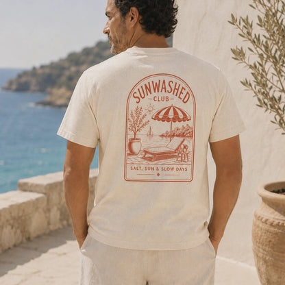 Man wearing a 'Sunwashed Club' t-shirt with a beach scene design, standing by a scenic coastal area.
