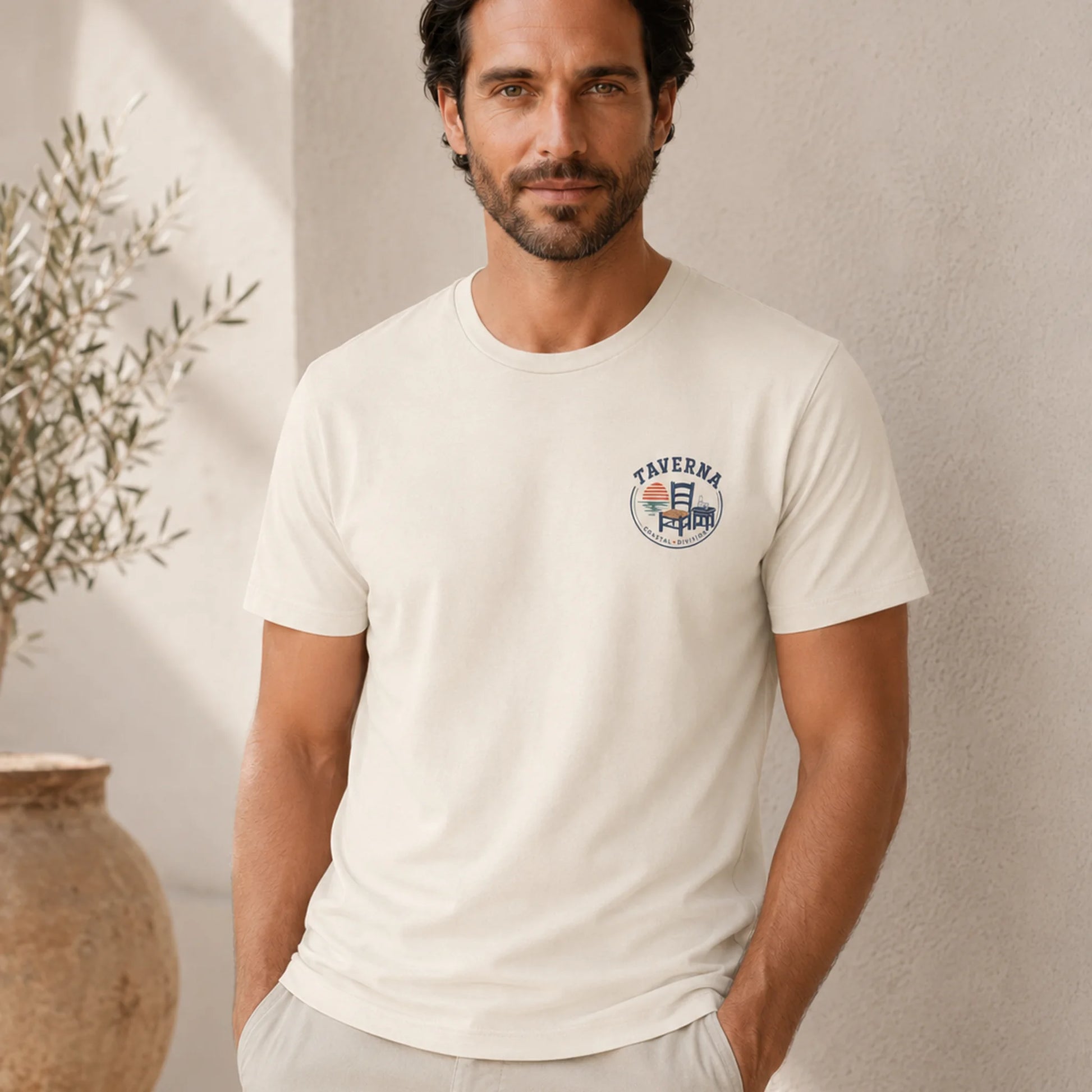Man wearing a white t-shirt with a logo, standing against a neutral wall with a plant.
