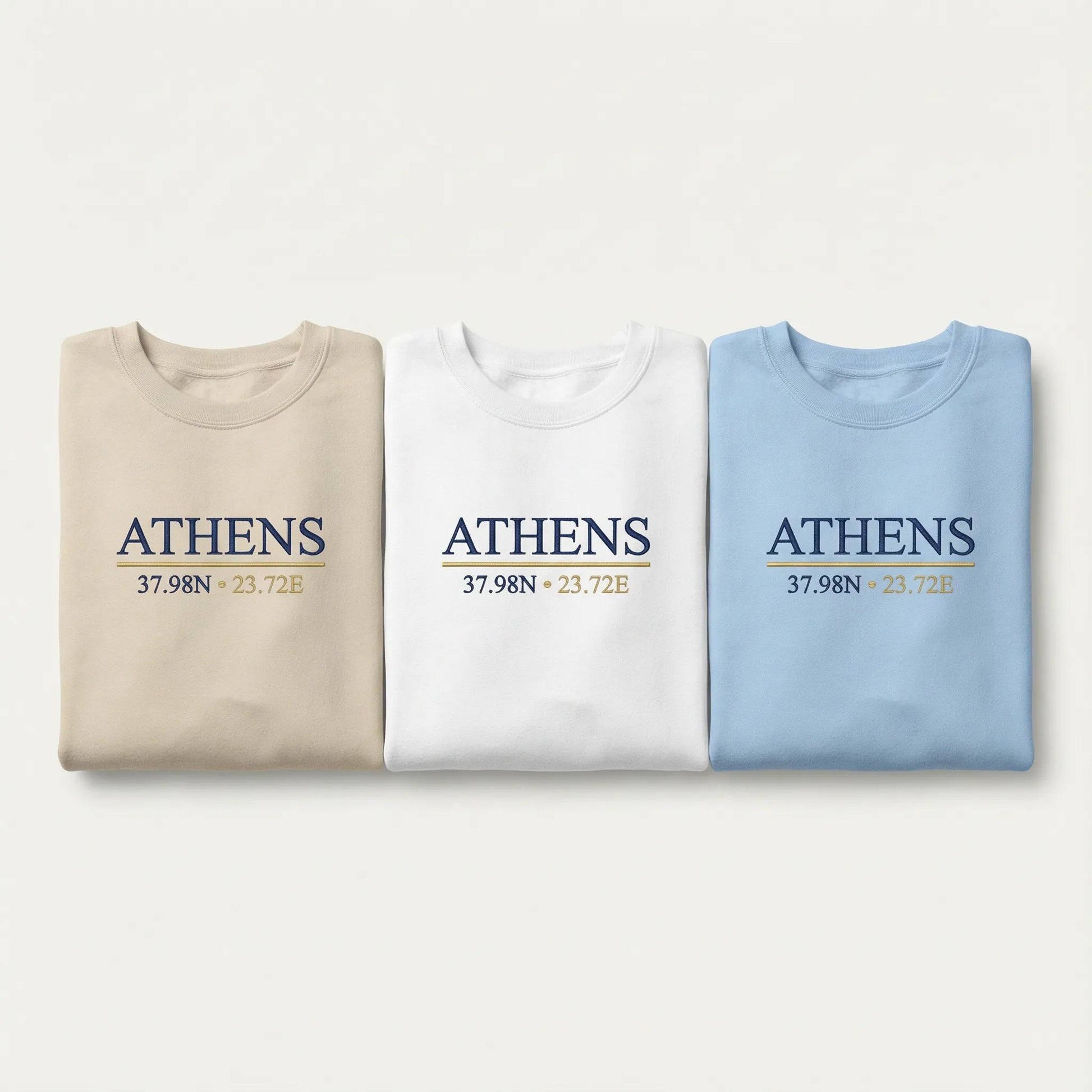 Athens coordinates embroidered crewneck sweatshirt color lineup showing Sand, White, and Light Blue options with navy and gold chest embroidery.