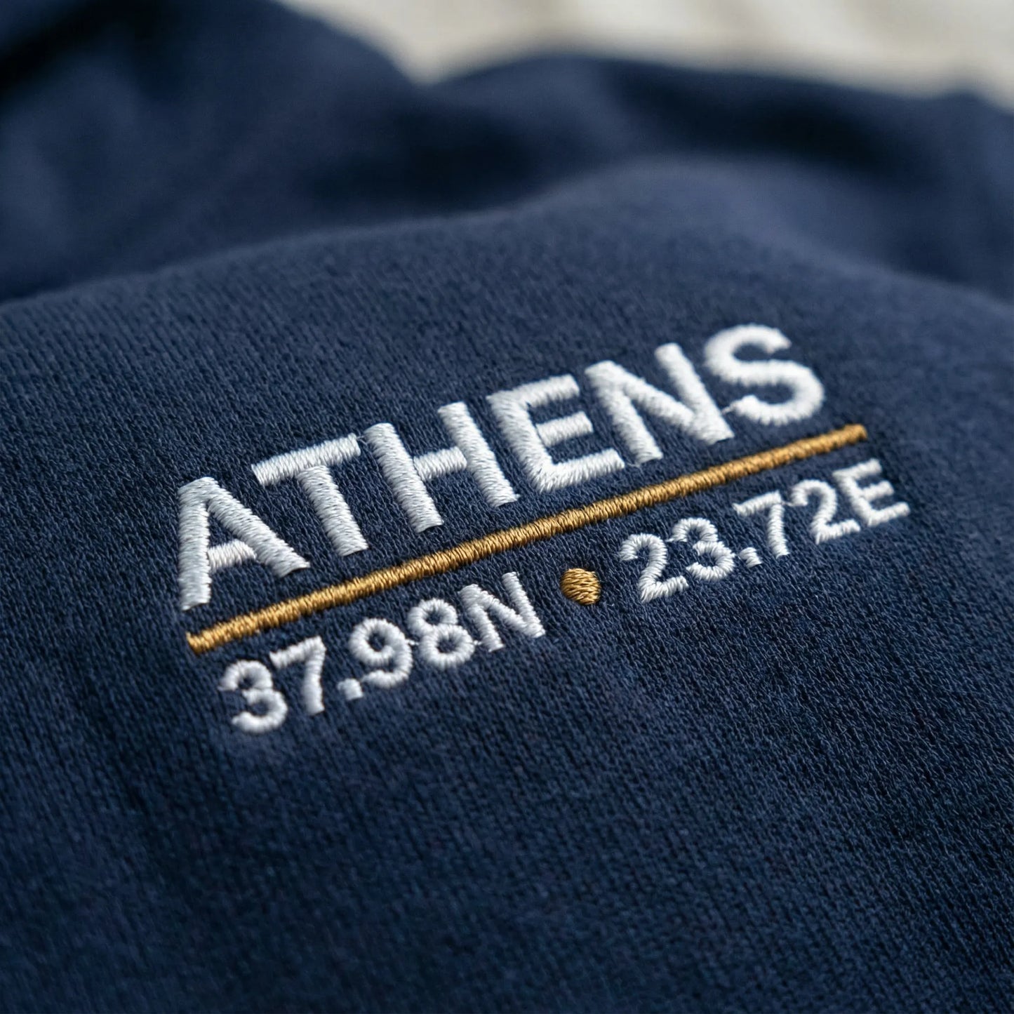 Macro close-up of Athens coordinates embroidery on navy hoodie left chest showing stitched ATHENS 37.98N 23.72E detail