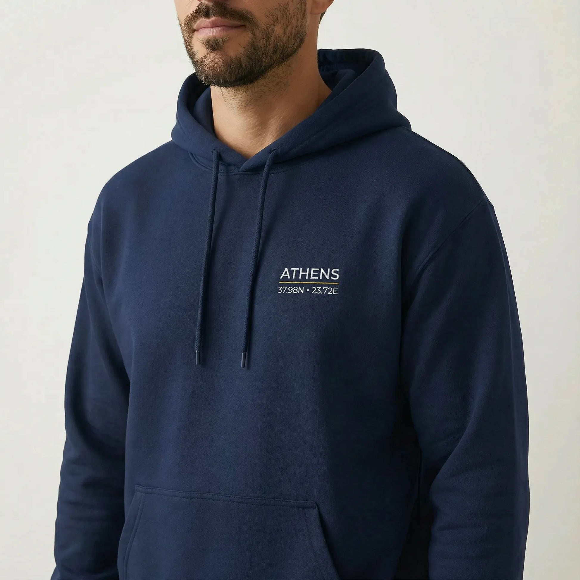 Close-up on model wearing navy hoodie with Athens coordinates left chest embroidery ATHENS 37.98N 23.72E visible