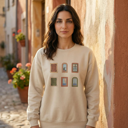 Woman wearing a beige sweatshirt with window designs, standing in a sunlit street.
