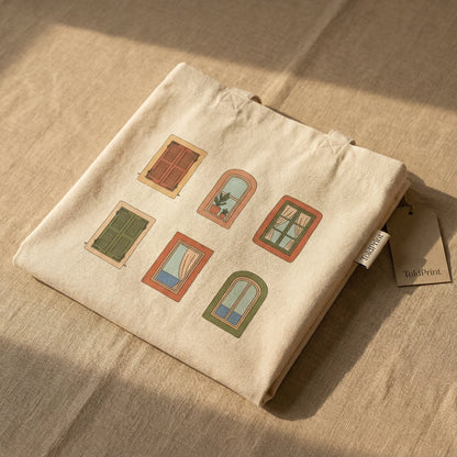 Photorealistic studio packaging shot of the Mediterranean Windows tote bag neatly folded on a neutral surface.