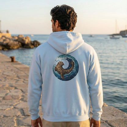 Unisex model wearing Monk Seal Embrace hoodie by the sea with back monk seal and wave design visible