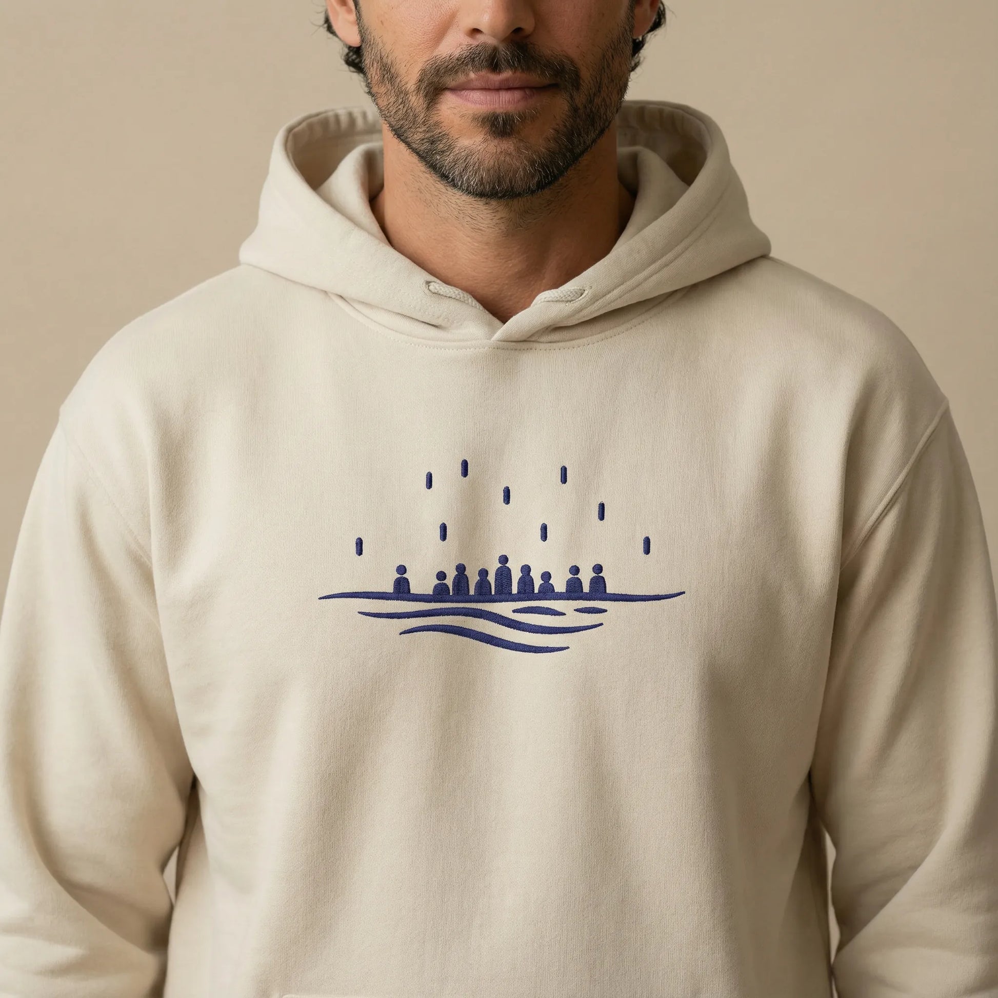 Close-up on model showing navy Sea Crossing embroidery centered on the chest of a sand hoodie.