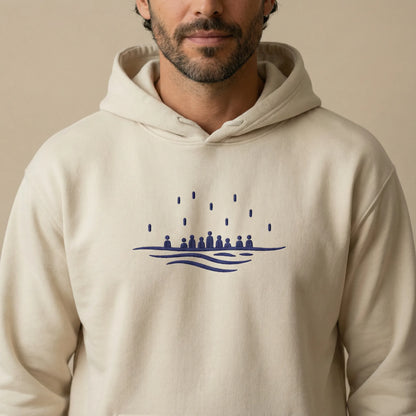 Close-up on model showing navy Sea Crossing embroidery centered on the chest of a sand hoodie.
