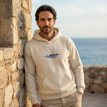 Outdoor Mediterranean shot of model wearing sand Sea Crossing hoodie with navy embroidered motif on the chest.