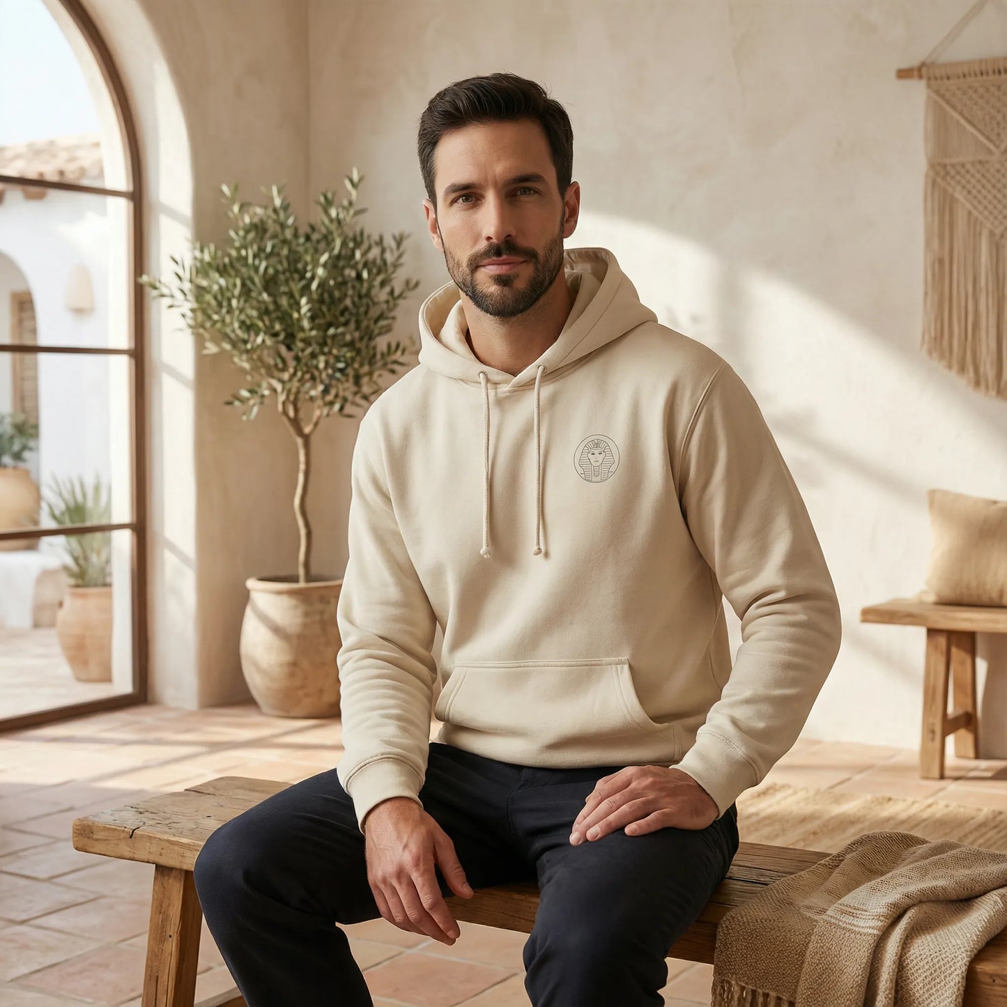 Unisex model indoors wearing sand Sphinx hoodie, front view with minimal chest badge and relaxed styling