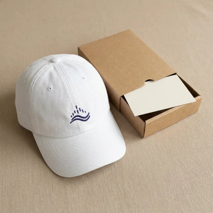 Packaging shot of white embroidered dad cap with navy wave emblem, minimalist unboxing scene on neutral beige background.