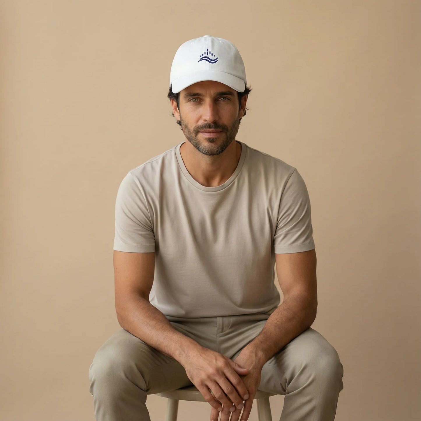 White embroidered dad cap on model, navy waves emblem, low profile unstructured cotton hat, studio lifestyle seated pose.