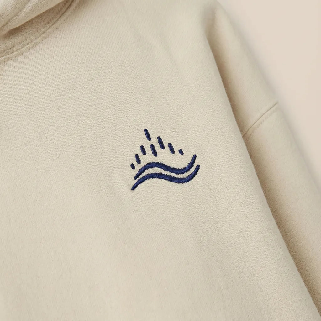Detail close-up of navy waves emblem embroidery on the left chest of a sand hoodie.