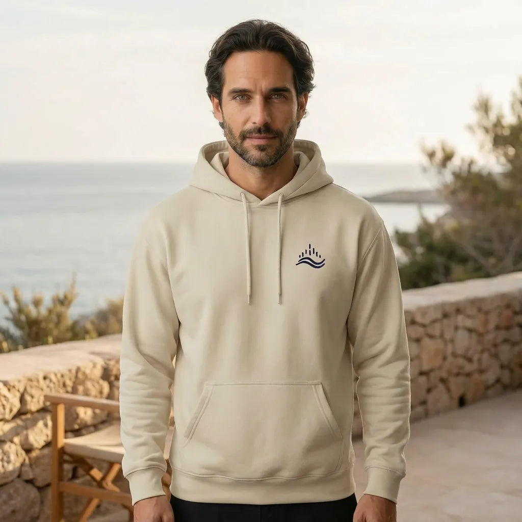 Outdoor Mediterranean lifestyle shot of a model in a sand hoodie with navy waves emblem on the left chest, standing by the sea.