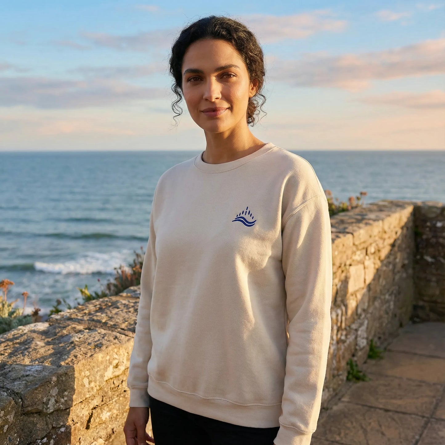 Outdoor Mediterranean scene with model wearing sand sweatshirt featuring navy waves emblem.