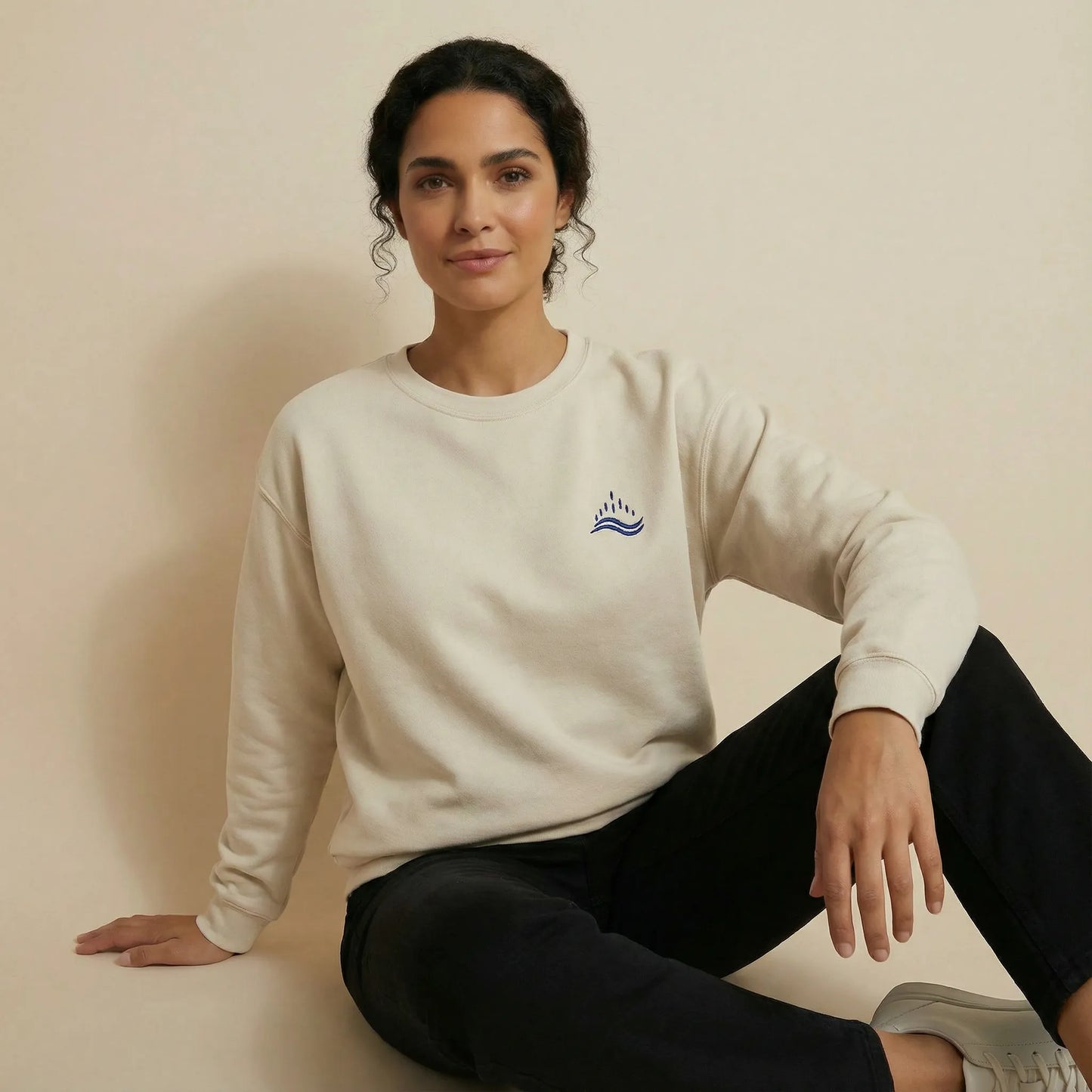 Studio lifestyle shot of a woman sitting relaxed on the floor wearing a sand sweatshirt with a small navy waves emblem embroidered on the left chest, minimalist Sea & Solidarity design.