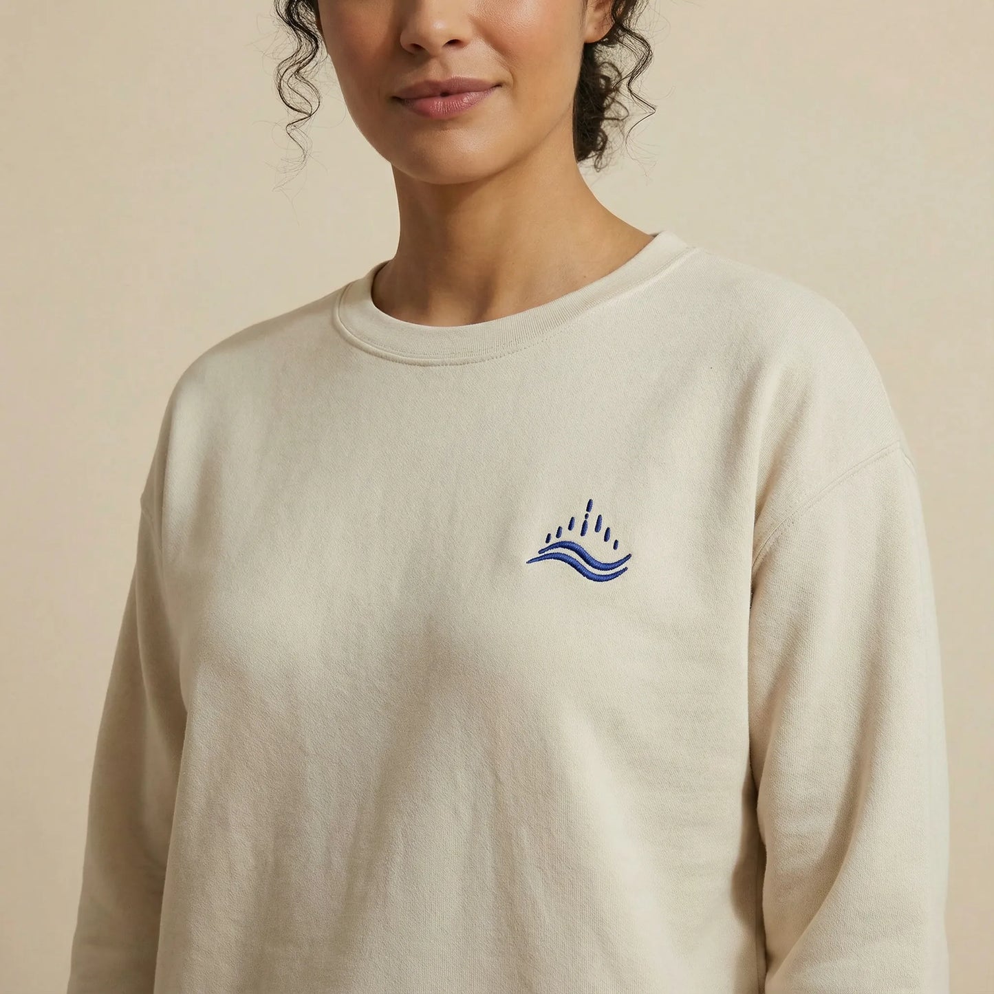 Close-up of the left chest on a sand unisex sweatshirt, showing the navy waves emblem embroidery symbolising sea and solidarity, soft minimalist crewneck fabric detail.