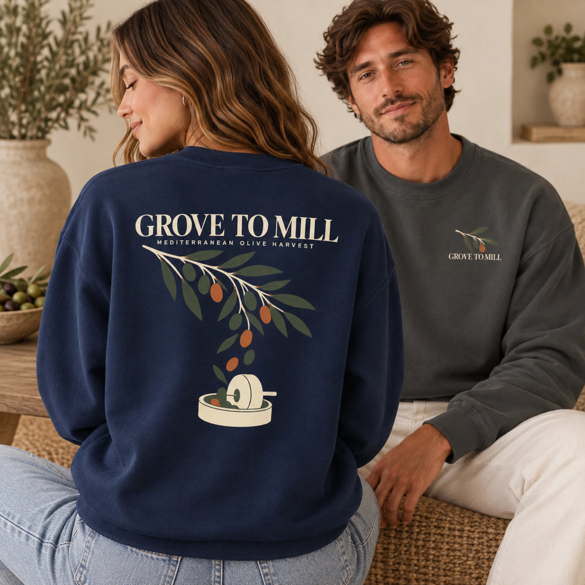 A man and a woman sitting indoors wearing the same crewneck sweatshirt from different angles. The woman in the foreground shows the back of the navy sweatshirt with a large olive harvest graphic and 'GROVE TO MILL' text, while the man in the background shows the front left-chest logo of the same sweatshirt.