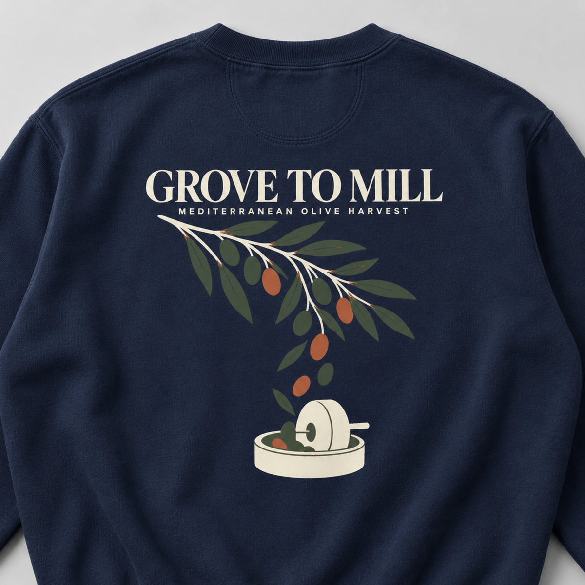 A navy blue crewneck sweatshirt featuring a graphic print of an olive branch with green leaves and orange olives, with a small mill illustration below. The text reads ' GROVE TO MILL' and 'MEDITERRANEAN OLIVE HARVEST'.