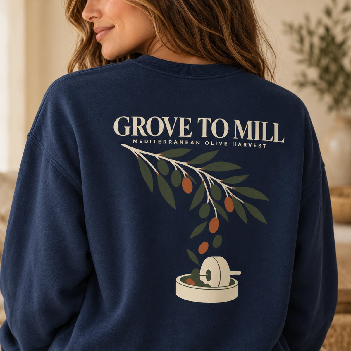 TP HERO: A woman wearing a navy blue sweatshirt, shown from the back. The sweatshirt features a graphic print of an olive branch with green leaves and small orange olives, with a milling wheel and the text 'GROVE TO MILL' and 'MEDITERRANEAN OLIVE HARVEST' in cream.