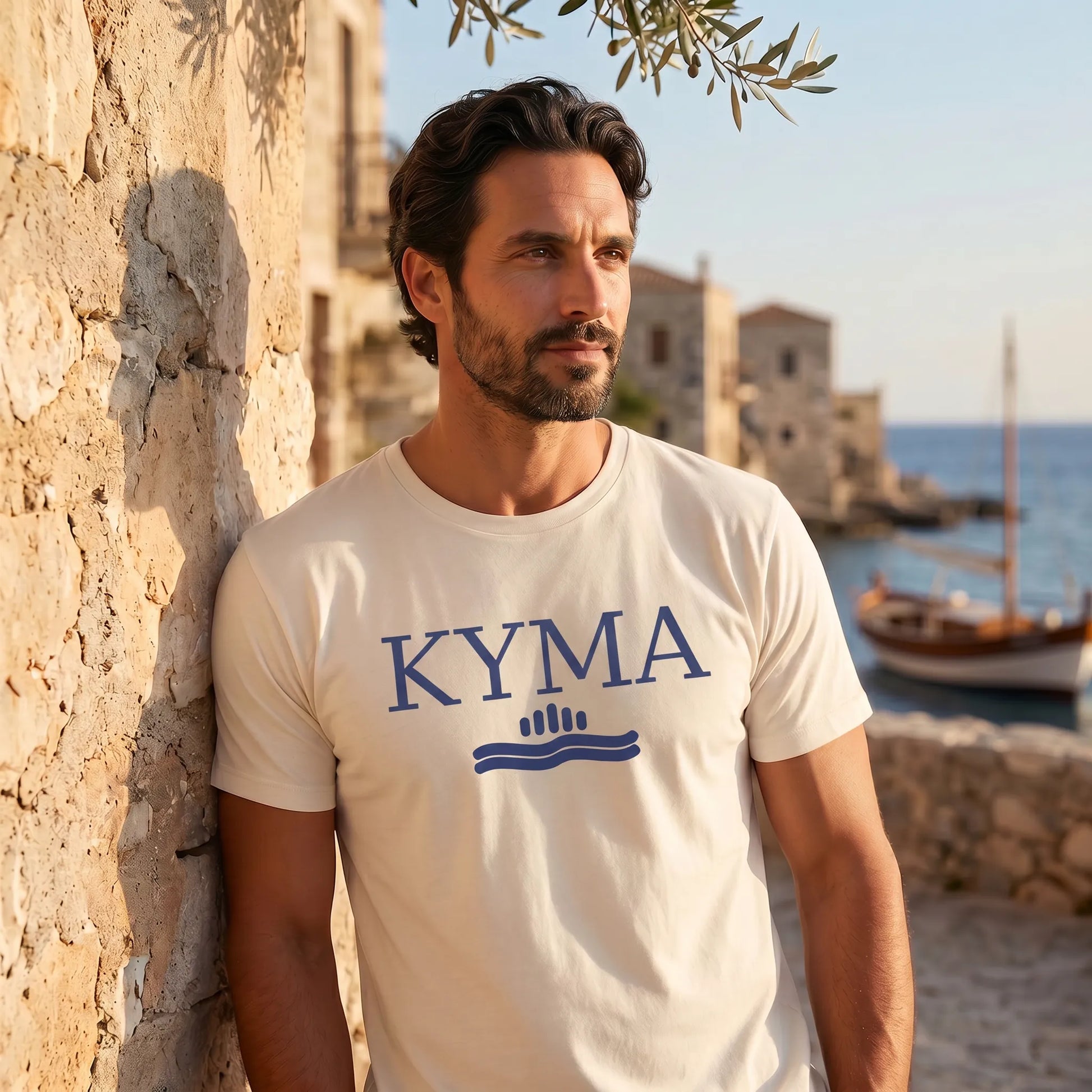 Man wearing a 'KYMA' t-shirt by a stone wall with a scenic background