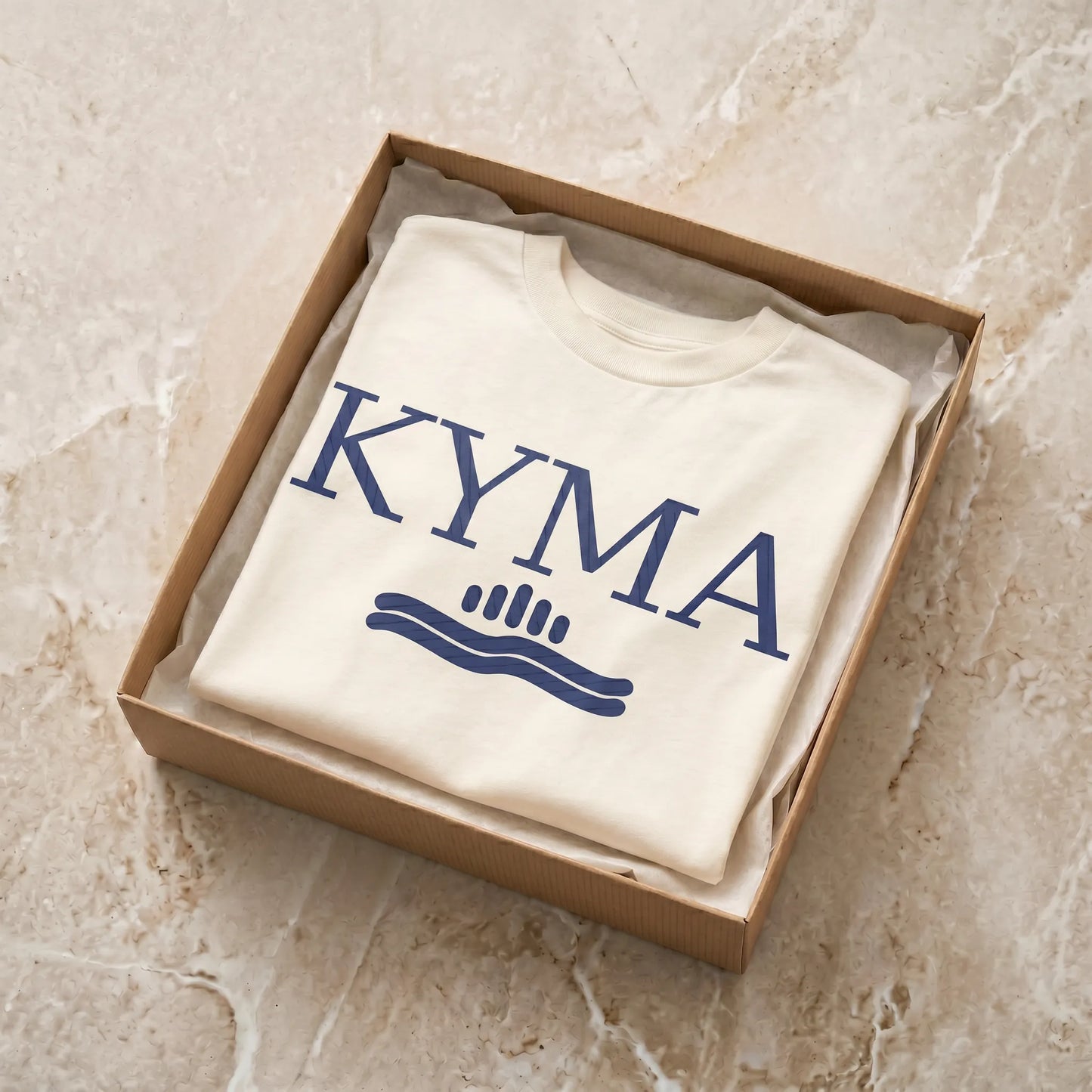 Folded white sweatshirt with 'KYMA' logo in a cardboard box on a beige marble surface