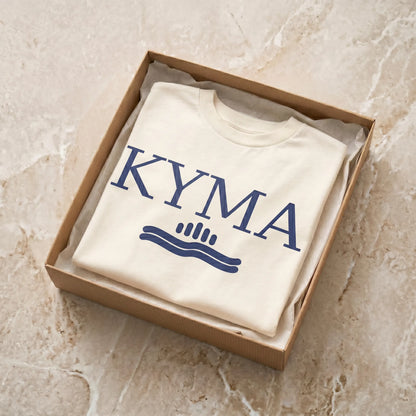 Folded white sweatshirt with 'KYMA' logo in a cardboard box on a beige marble surface