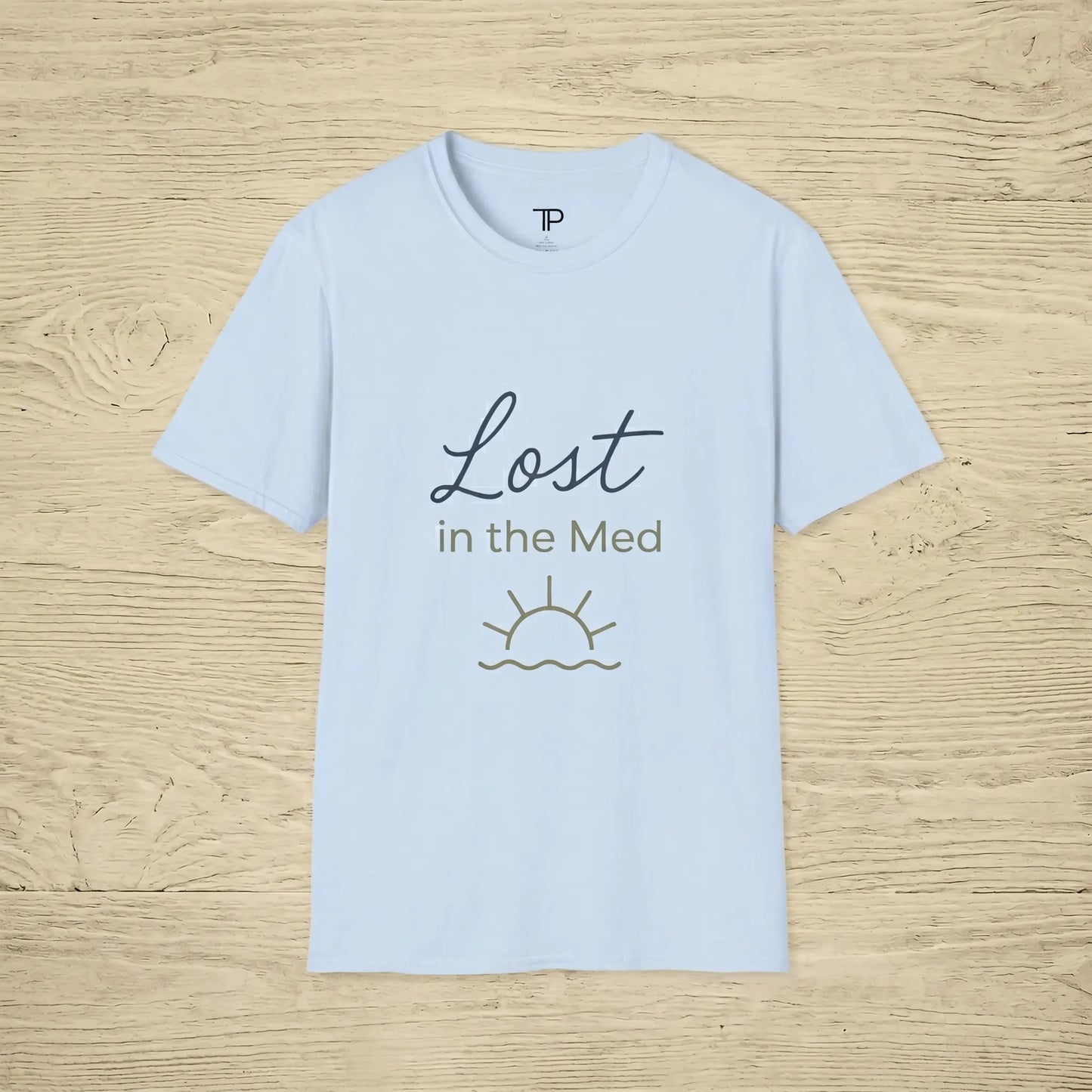Light blue t-shirt with 'Lost in the Med' text and sun graphic on a wooden surface