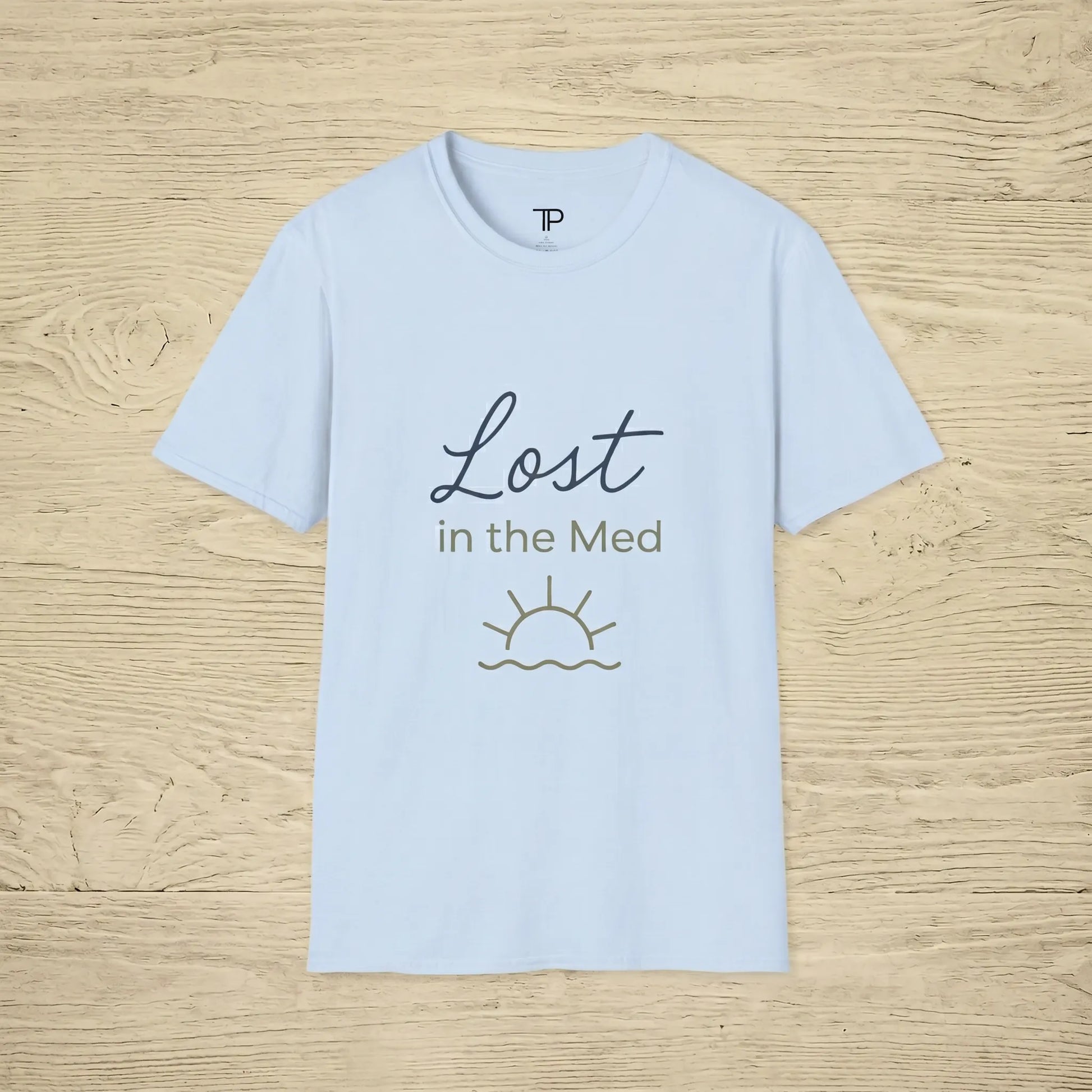 Light blue t-shirt with 'Lost in the Med' text and sun graphic on a wooden surface