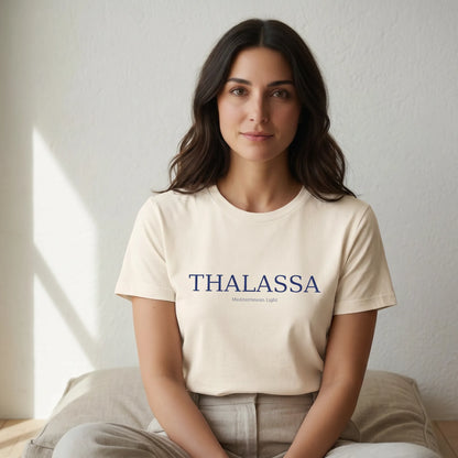 Woman wearing a beige t-shirt with 'THALASSA' text sitting on a bed.