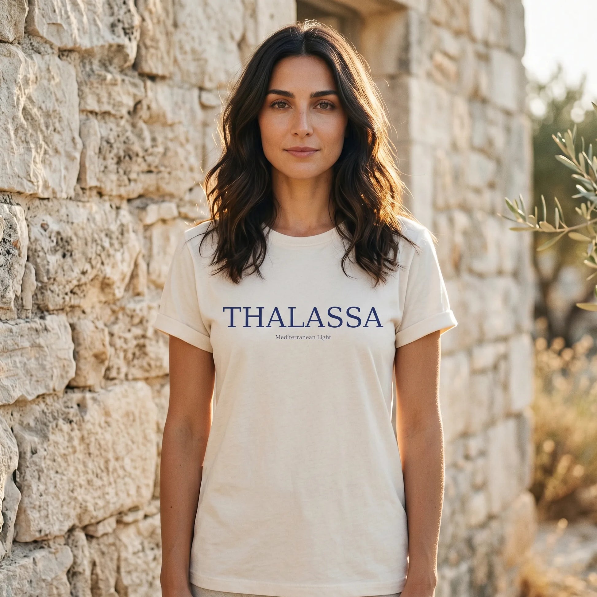Woman wearing a 'Thalassa' t-shirt standing in front of a stone wall.